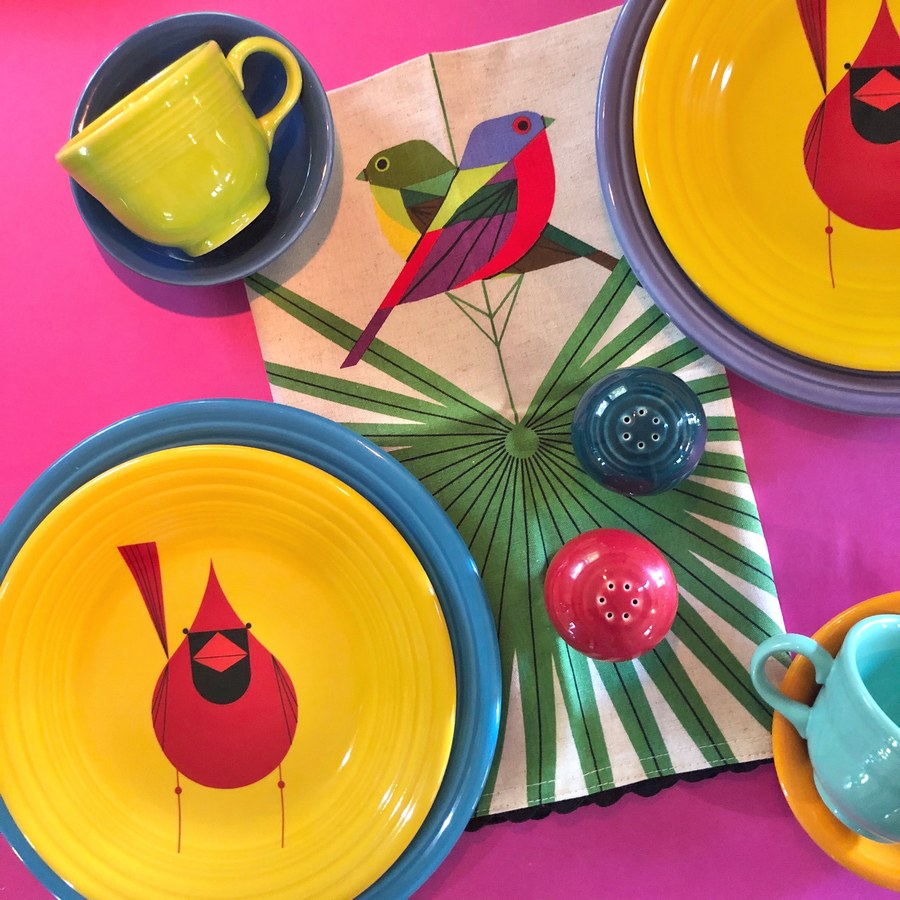 Fiesta plates arranged with a Flamboyant Feathers tea-towel on a pink background, with brightly colored mugs and saucers nearby